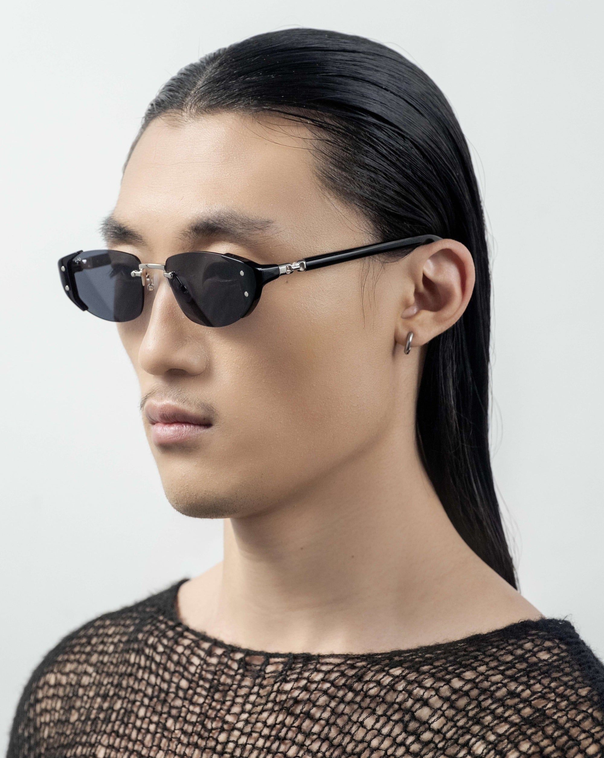 A person with long, sleek black hair and wearing For Art's Sake® Harbour sunglasses with a retro-inspired design looks to the side. They have a small hoop earring in one ear and are dressed in a textured, black mesh top. The background is plain white.