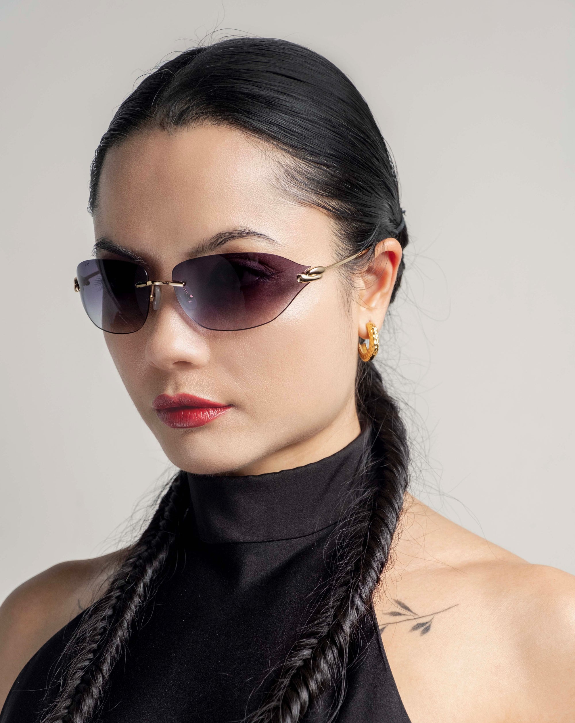 A woman with long, dark braided hair and 18-karat gold hoop earrings wears For Art's Sake® Serpent I sunglasses featuring a bold frameless wrap lens design. She has a tattoo near her shoulder and is looking confidently into the camera against a neutral background in her sleeveless black top.