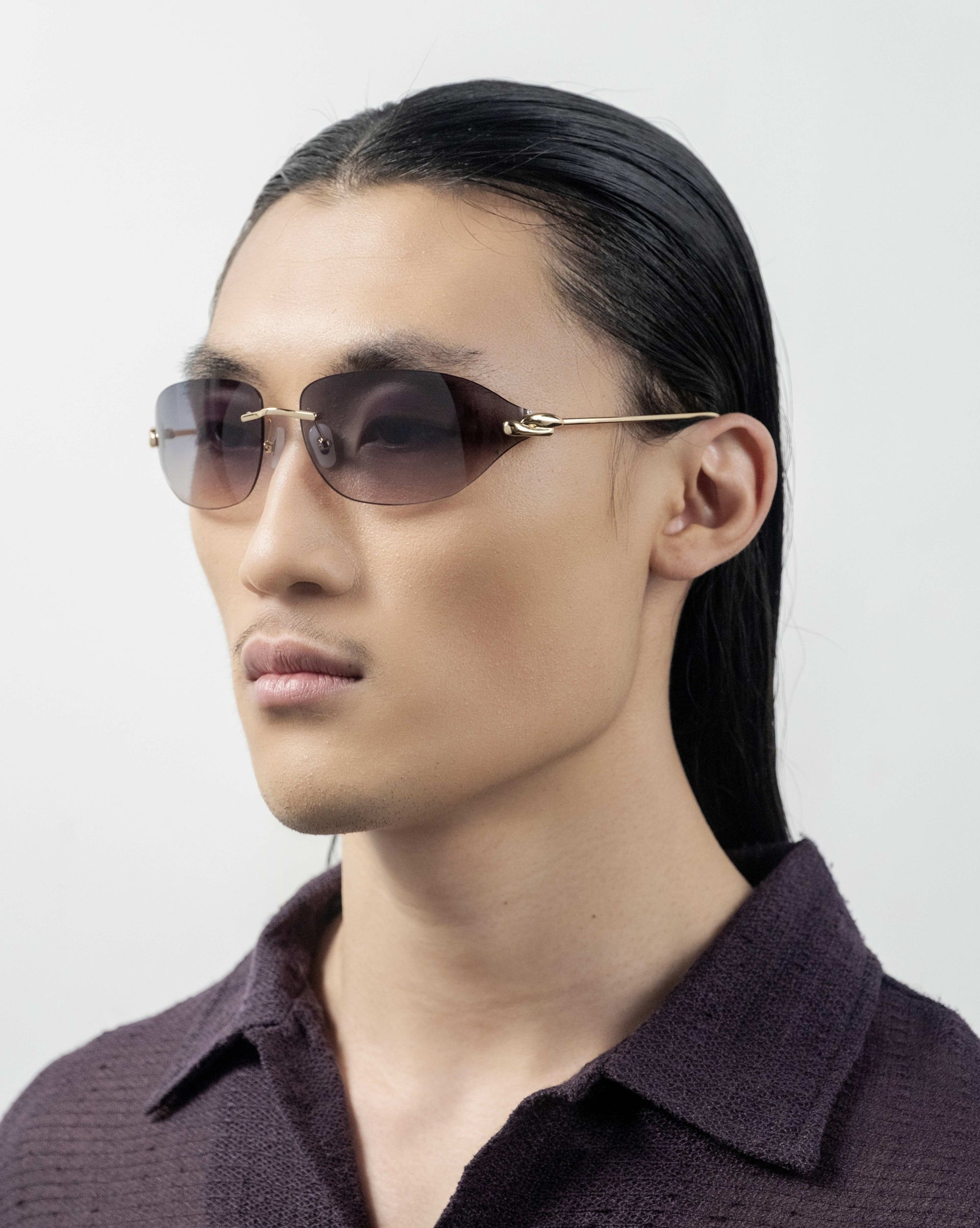 A person with long, sleek dark hair is seen wearing For Art's Sake® Serpent I frameless wrap lens oval-shaped sunglasses with golden temple arms. They are dressed in a dark, textured shirt and facing slightly left against a plain, light background.