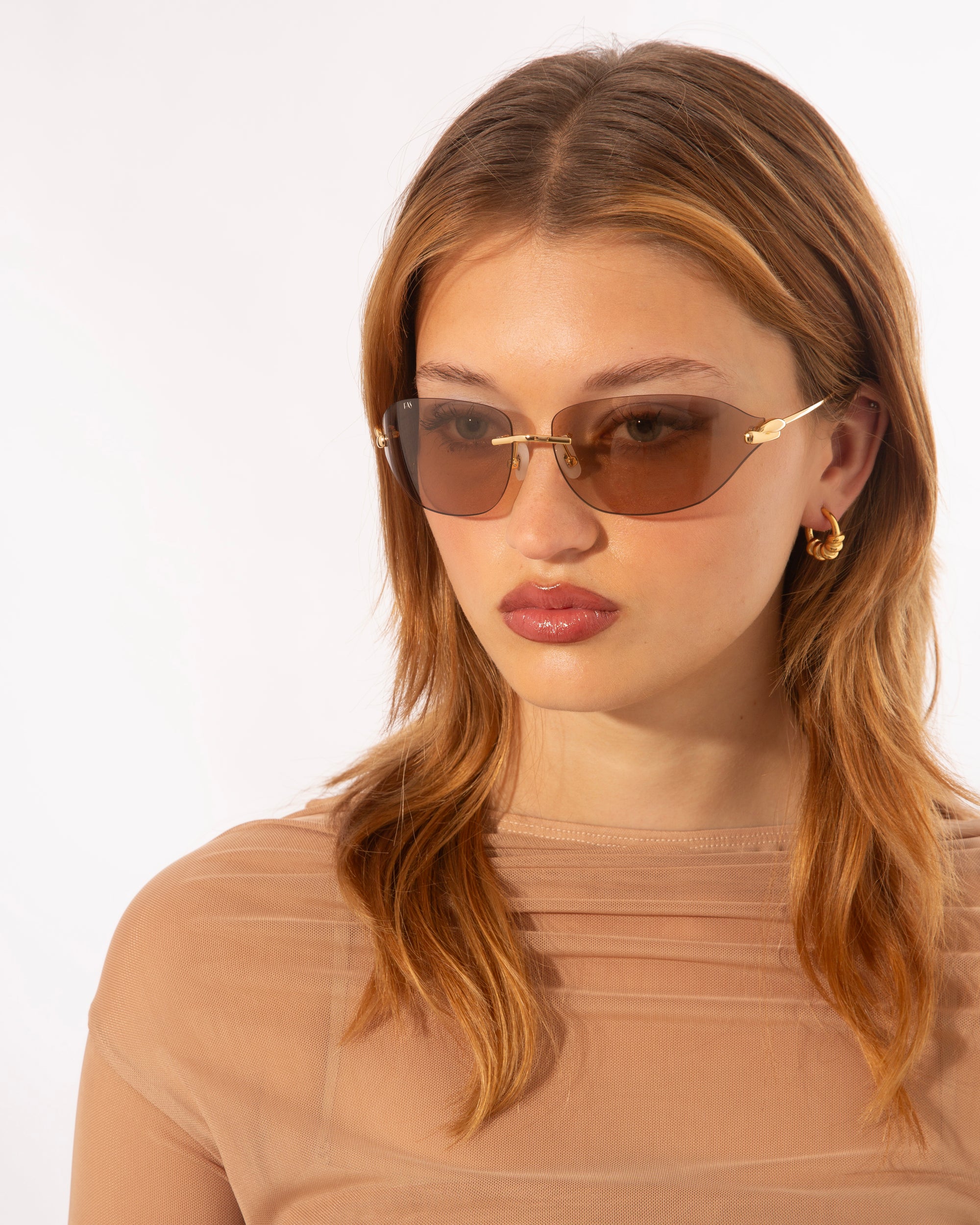 A person with long brown hair wearing For Art's Sake® Serpent I sunglasses and gold hoop earrings. They are dressed in a translucent beige top and look slightly to the side against a plain white background.
