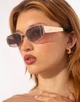 A woman with light brown hair models For Art's Sake® Talia geometric gold-plated sunglasses with dark lenses and gold hoop earrings. She wears natural makeup and looks to the side against a plain background.