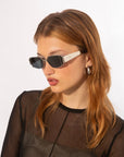 A woman with light brown hair wears the Talia geometric sunglasses by For Art's Sake®, featuring white metal frames, along with silver hoop earrings and a sheer black top, posing against a plain white background.