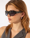 A woman with light brown hair wears For Art's Sake® Raven sunglasses, a sleeveless gray top, and silver drop earrings, looking to the left against a plain white background.