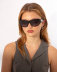 A woman with light brown hair poses against a plain white background wearing oversized For Art's Sake® Raven sunglasses, silver earrings, and a gray sleeveless collared top.