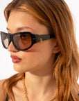 A woman with light brown hair wears oversized For Art's Sake® Raven sunglasses with brown lenses and an 18-karat gold chain necklace, gazing slightly to the side against a plain background.