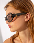 A woman with light skin and auburn hair wears For Art's Sake® Raven sunglasses featuring a small gold hoop detail and an 18-karat gold chain necklace, shown in profile against a plain background.