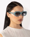 A person with long, straight black hair is wearing blue, hexagonal sunglasses from For Art's Sake® and a white, sleeveless top. The retro glamour of their look is accentuated by the geometric Vitamin D sunglasses against the plain white background.