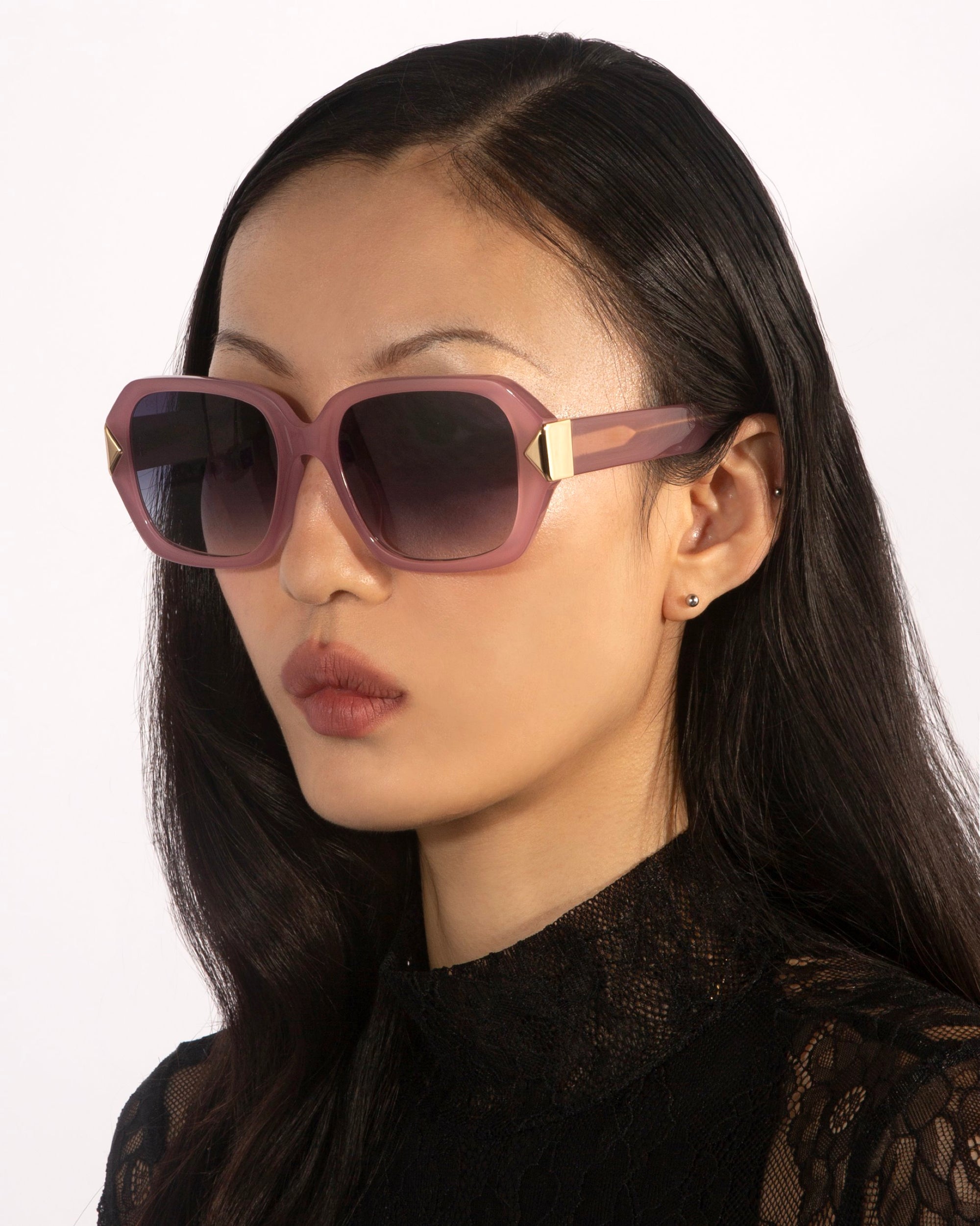 A woman with long dark hair wears For Art's Sake® Frankie Plum oversized geometric sunglasses with gold accents and a black lace top, gazing to the side against a plain light background.