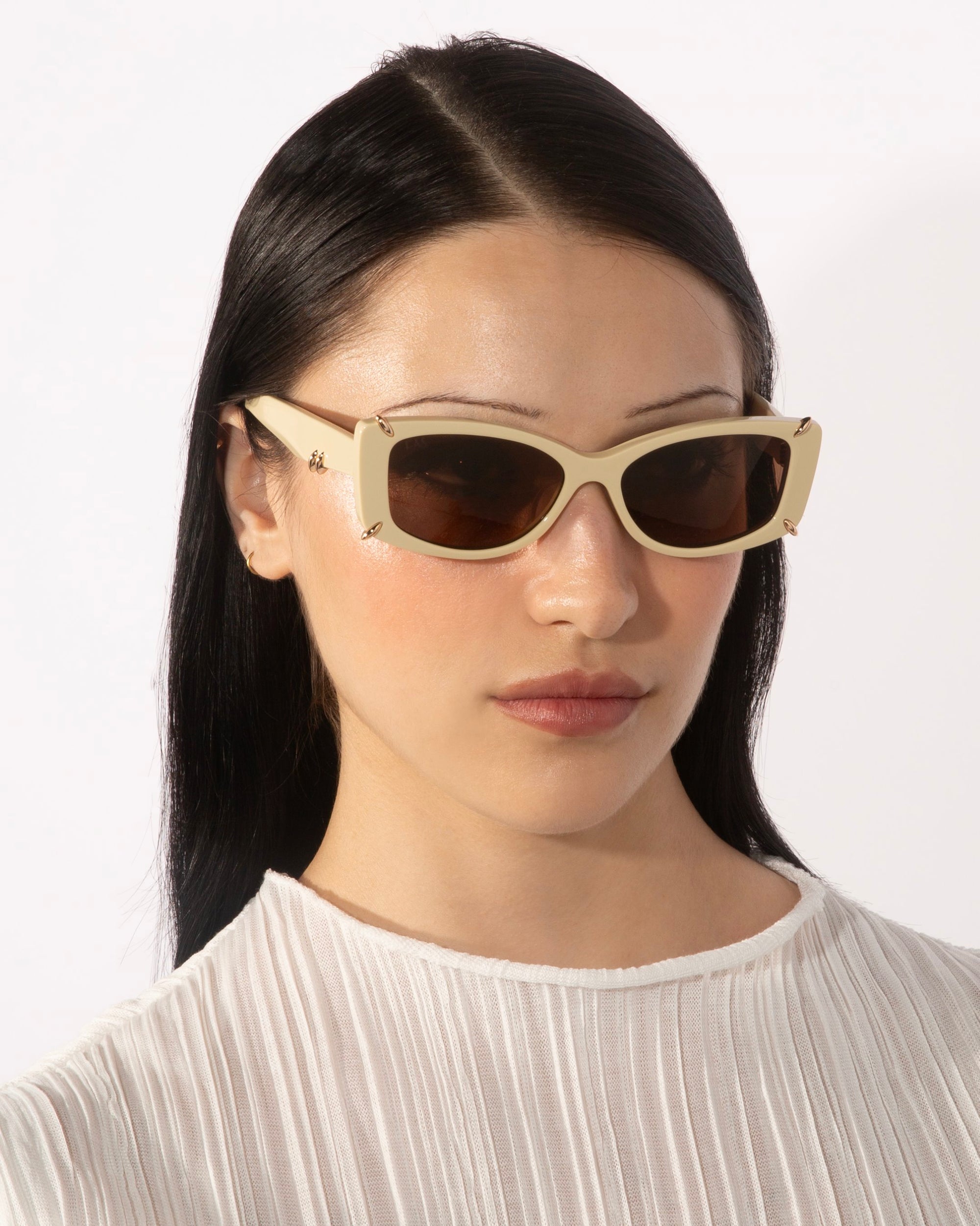 A person with long black hair wears For Art's Sake® Dyer rectangular beige luxury sunglasses with dark lenses and a white, pleated blouse. The background is plain white.