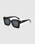 The Rectangular black Ribbon sunglasses from For Art's Sake® boast dark lenses and a thick, black frame. This chic accessory is enhanced with a silver decorative element near the hinges. The sustainable sunglasses are positioned at an angle to the left against a gray background.