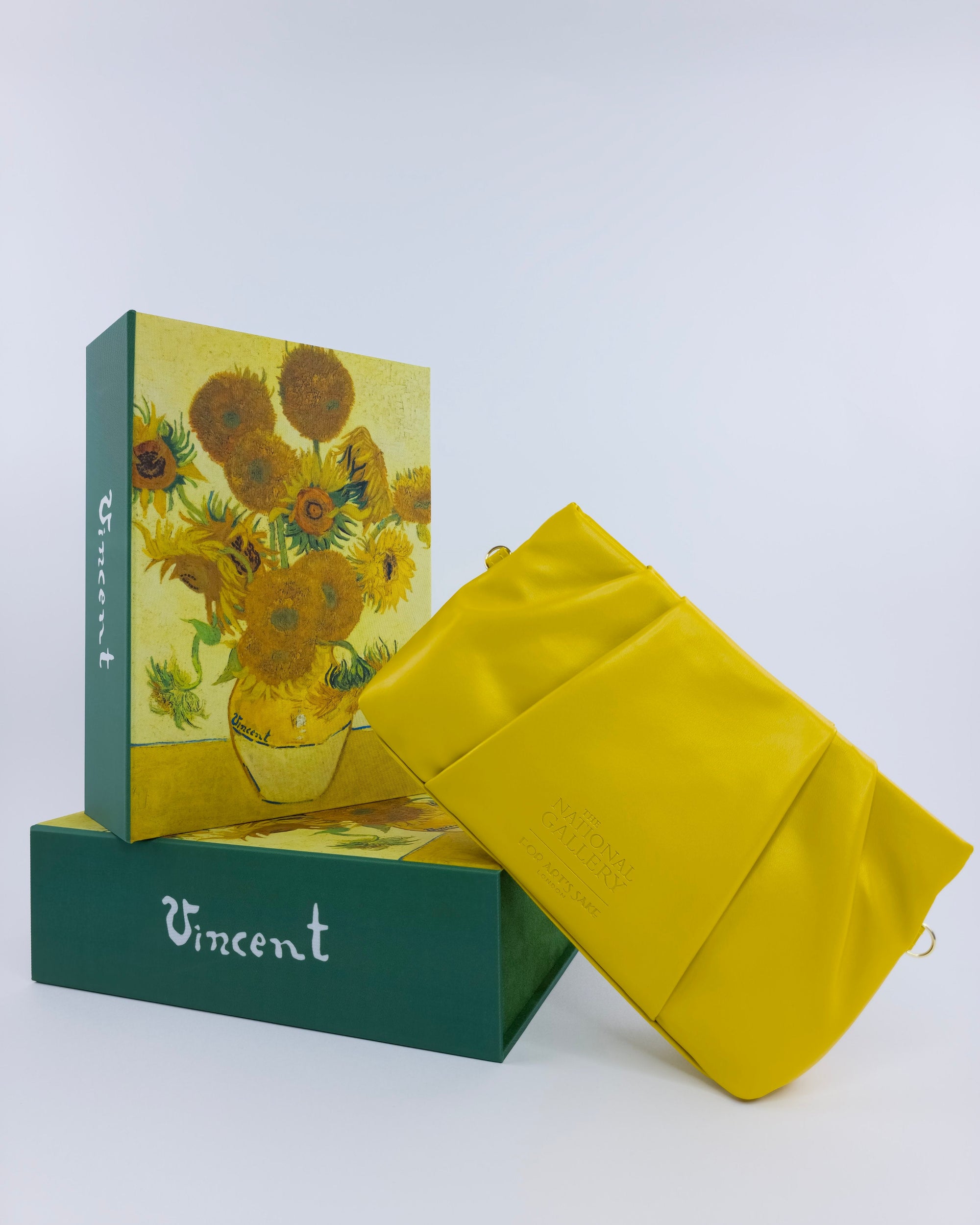 A yellow clutch sits on two green boxes featuring Vincent van Gogh’s "Sunflowers" and name, perfectly displaying the Vincent Van Gogh sunglasses by For Art's Sake® against a plain white background.