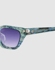 Close-up of Claude Monet (Limited Edition) cat-eye sunglasses by For Art's Sake®, featuring a blue and green marbled frame, purple tinted lenses, and gold text on the arm.