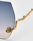 The La Vie En Rose sunglasses from For Art's Sake® feature a gradient lens transitioning from blue to clear. The gold frame, designed like branches, is embellished with sculpted rose ornaments for a floral touch of elegance.