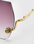 Check out the La Vie En Rose eyeglass frame by For Art's Sake®, part of the Rose Garden Collection. It features oversized frameless lenses with a purple-to-clear gradient, and a gold arm adorned with sculpted rose ornaments at the hinge, leading to a wavy design.