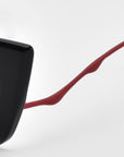 A close-up showcases Dusk by For Art's Sake®: a sleek black object with a glossy finish, adorned with a long, irregularly curved red appendage that resembles a cat-eye silhouette. This is set against a neutral gray background, enhancing its sophisticated vibe.