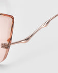Close-up of For Art's Sake® Dusk eyeglasses featuring pink-tinted lenses and thin rose-gold frames. The sophisticated design showcases a unique waved shape on the temple arm against a plain background.