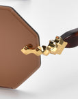 Close-up of the Haze glasses by For Art's Sake® with a distinctive gold geometric temple design and intricate decorations. The lenses are angular brown hexagons on a plain white background.
