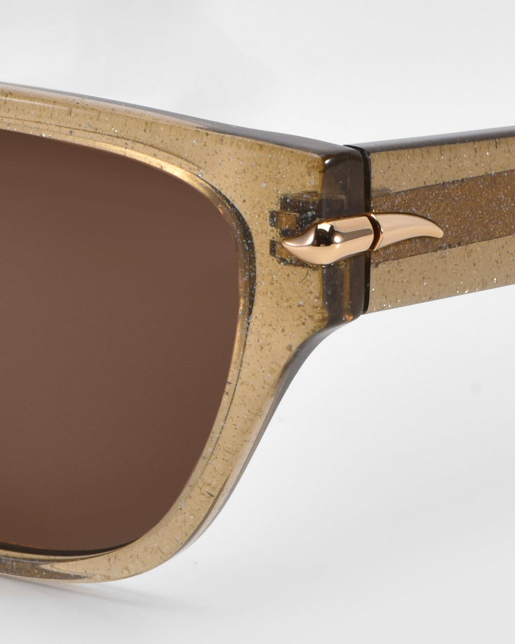 Close-up of For Art's Sake® Rosie everyday sunglasses with glittery, translucent brown frames and dark brown lenses. The cat-eye acetate hinge features a gold metallic accent like an abstract bird's head on a light gray background.
