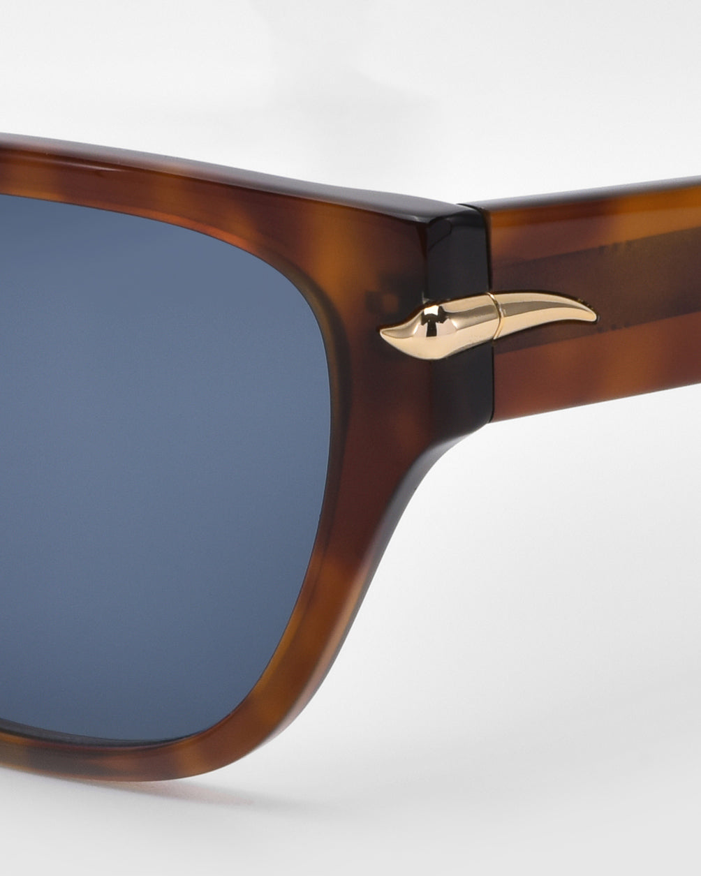 Close-up of For Art's Sake® Rosie tortoiseshell sunglasses featuring blue lenses and a gold emblem on the temple hinge, set against a white background.