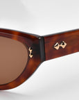 Close-up of For Art's Sake® Lush sunglasses in a brown cat-eye style, with brass bow tie-shaped studs near the hinge and dark-tinted lenses. The premium tortoiseshell-patterned frame stands out against a minimalist white background.
