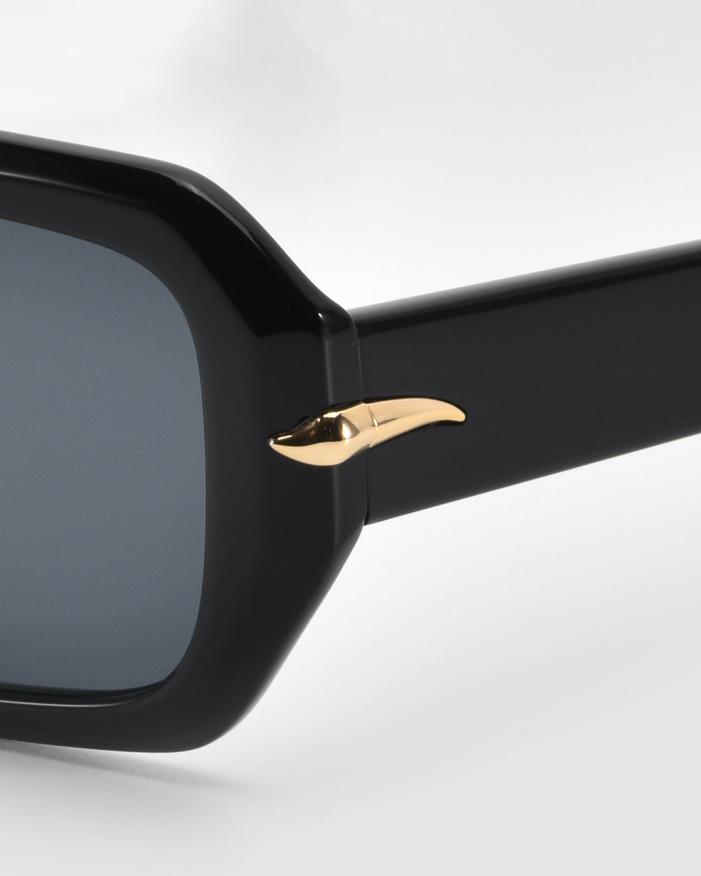 Close-up of For Art's Sake® Fleur sunglasses in black, featuring a minimalist design with a gold metallic accent shaped like a small curved horn on the temple. The dark rectangular lens stands out against a plain light background.