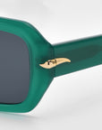 A close-up of the For Art's Sake® Fleur sunglasses shows a minimalist green frame with tinted lenses. The temple hinge features a gold metallic accent resembling a horn or curved spike, set against a white background.