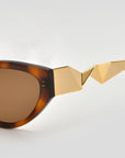 Close-up of For Art’s Sake® Unfold sunglasses featuring brown lenses and ornate gold geometric details on pleated temples, set against a plain light background.