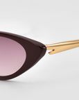 Close-up of For Art's Sake® Slash cat-eye sunglasses with a dark burgundy frame, pink-tinted lens, and gold metal arm, highlighting sculpted details against a light gray background.