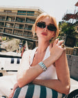A red-haired woman in black Lucio sunglasses by For Art’s Sake®, a white tank top, and silver jewelry sits on a striped towel in a boat with palm trees and a modern building in the sunny background.