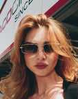 A woman with long red hair wears For Art’s Sake® Lucio sunglasses outside, with a building and an "ACO" sign in the background.