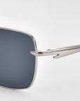 Close-up of the left lens and arm of For Art's Sake® Edge sunglasses, featuring a sleek square silhouette with silver metal frames and dark lenses against a plain light background.