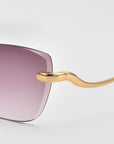 Close-up of For Art's Sake® Spark rimless cat-eye sunglasses featuring a gold arm and a gradient lens fading from purple to clear, set against a plain light gray background.