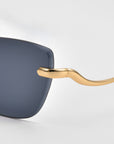 Close-up of For Art's Sake® Spark rimless cat-eye sunglasses with a gold frame and dark tinted lens, highlighting the smooth, curved temple design against a light gray background.
