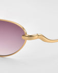 Close-up of For Art's Sake® Rebirth sunglasses with a sculptural gold metal frame, wavy temples, and purple-tinted lenses, set against a light gray background—a timeless statement accessory.