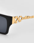 Close-up of For Art's Sake® Incomplete black square sunglasses with distinctive geometric mosaic metal arms, shown on a plain light gray background.