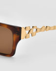 Close-up of For Art's Sake® Incomplete square sunglasses featuring brown acetate frames and gold geometric mosaic metal temples, set against a plain light gray background.