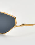 Close-up of For Art's Sake® Moment sunglasses, featuring a shiny gold metal frame and arms with dark geometric lenses, set against a plain light background.