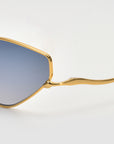 Close-up of For Art's Sake® Moment sunglasses, showcasing a gold geometric frame and blue-tinted lenses against a light background, with only part of the lens and frame in view.