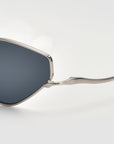 Close-up of For Art's Sake® Moment sunglasses with a geometric silhouette and silver metal frame, set against a light gray background. The photo shows part of one dark tinted lens and the temple arm.