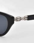 Close-up of For Art's Sake® "The Cuts" black cat-eye sunglasses with a distinctive silver metal chain linking the front frame to the temple arm, set against a plain light gray background.