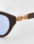 Close-up of The Cuts by For Art's Sake®—sculptural sunglasses with a weaved metal frame, brown arms, blue-tinted lenses, and gold chain-like temple details, set against a light gray background.