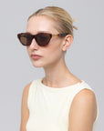A woman with blonde hair pulled back models The Cuts cat-eye sunglasses by For Art's Sake®, paired with a sleeveless cream top against a plain light gray background.