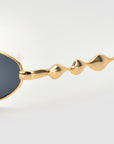 Close-up of For Art's Sake® Droplets sunglasses: gold, wavy arms and dark oval lenses offer a bold, retro statement against a simple light background.