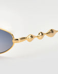 Close-up of Droplets by For Art's Sake®—oval sunglasses with gold wavy temples and blue-tinted lenses—set against a plain light background.