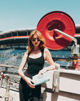 A woman in a black tank top and For Art's Sake® Droplets retro sunglasses stands by a red industrial reel at an outdoor stadium, holding a light blue clutch with empty bleachers behind her.