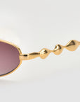 Close-up of For Art's Sake® Droplets: gold-framed oval sunglasses with wavy arms and purple-tinted lenses, set against a light grey background.