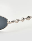 Close-up of For Art's Sake® Droplets: silver sunglasses with dark lenses and wavy, sculptural arms, ideal for bold, retro style lovers, set against a plain light gray background.