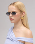 A woman with straight blonde hair wears For Art's Sake® Calla I lightweight sculptural eyewear, gold geometric earrings, and a light blue off-the-shoulder top, facing slightly to the side against a plain background.