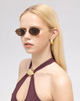 A woman with long blonde hair wears For Art's Sake® Calla I modern oval metal eyewear, gold earrings, and a dark halter-neck top with a round gold clasp, standing against a plain light background.