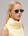 A woman with straight blonde hair wears For Art's Sake® Calla I oval metal silhouette sunglasses, gold earrings, and a cream sleeveless high-collar top. She faces slightly to the side against a plain background.